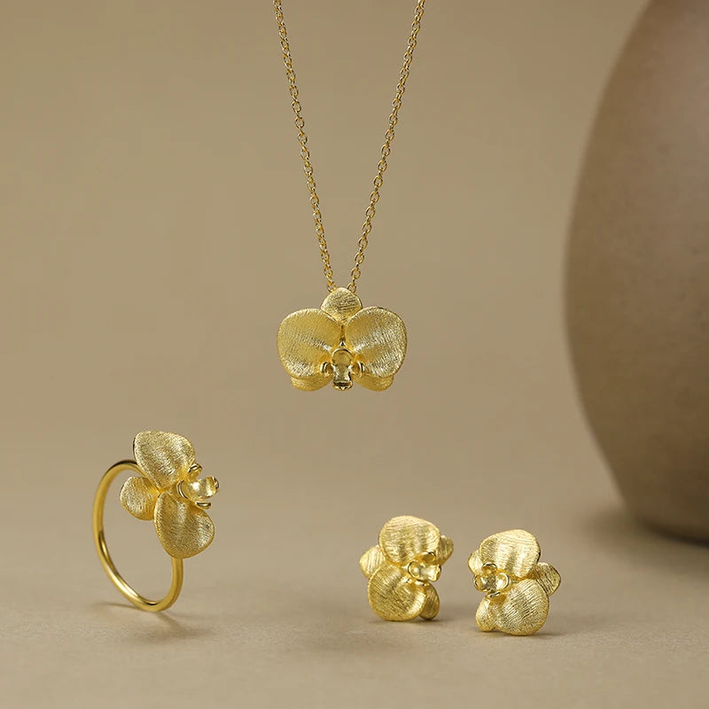 Gold orchid jewelry set featuring a necklace, ring, and stud earrings with textured flower details, displayed on a warm beige background.