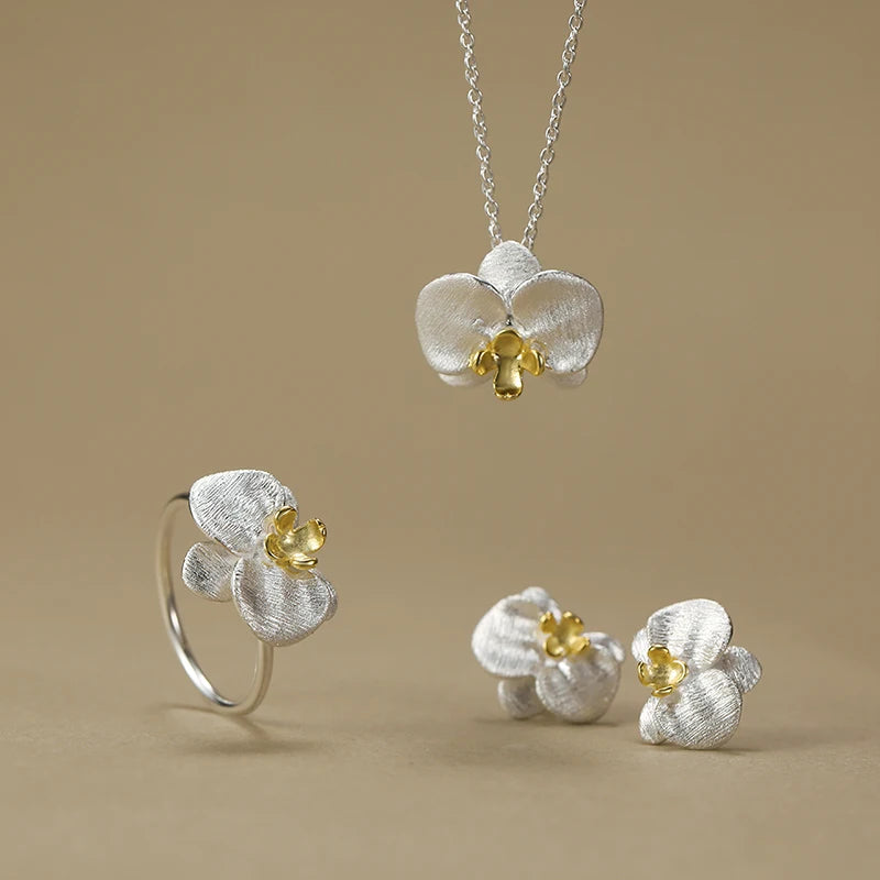 A jewelry set featuring a silver orchid pendant necklace, matching orchid ring, and orchid stud earrings, all with textured silver petals and yellow flower centers, displayed on a warm beige background.