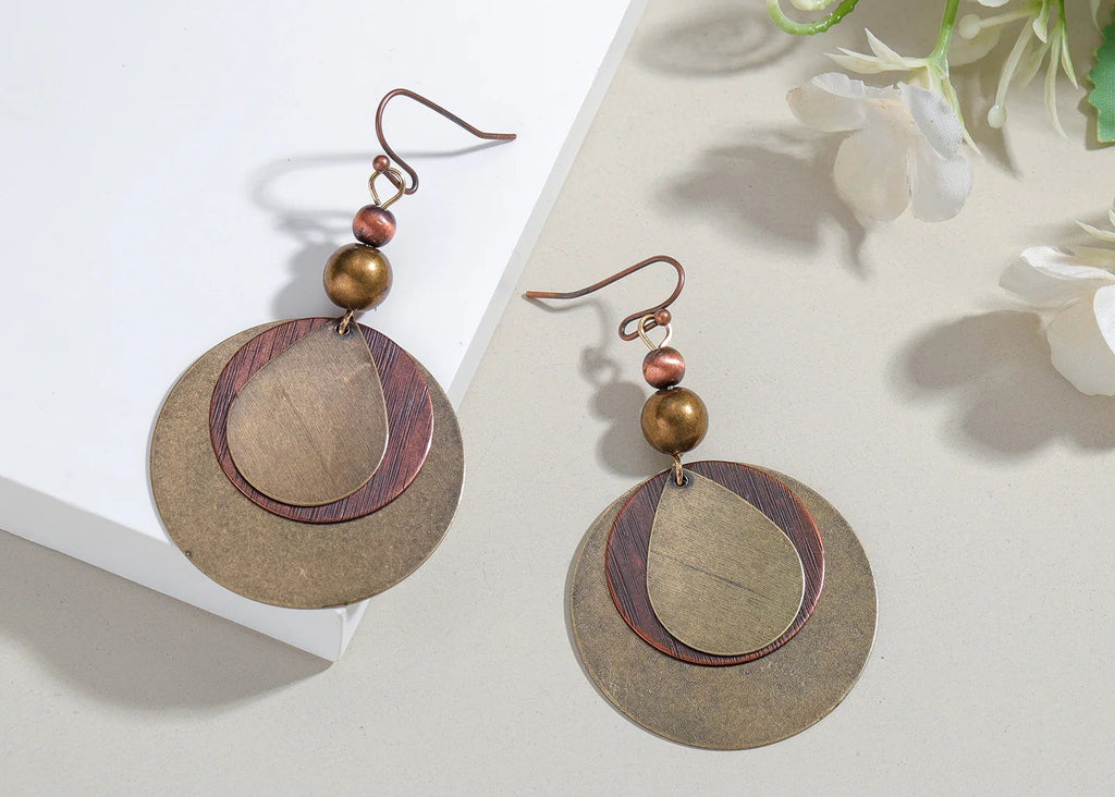 A pair of handmade dangle earrings with circular bronze-toned metal discs, a smaller concentric oval with a dark wood inlay, and a small round bead, displayed on a light beige surface next to a white box and white flowers.